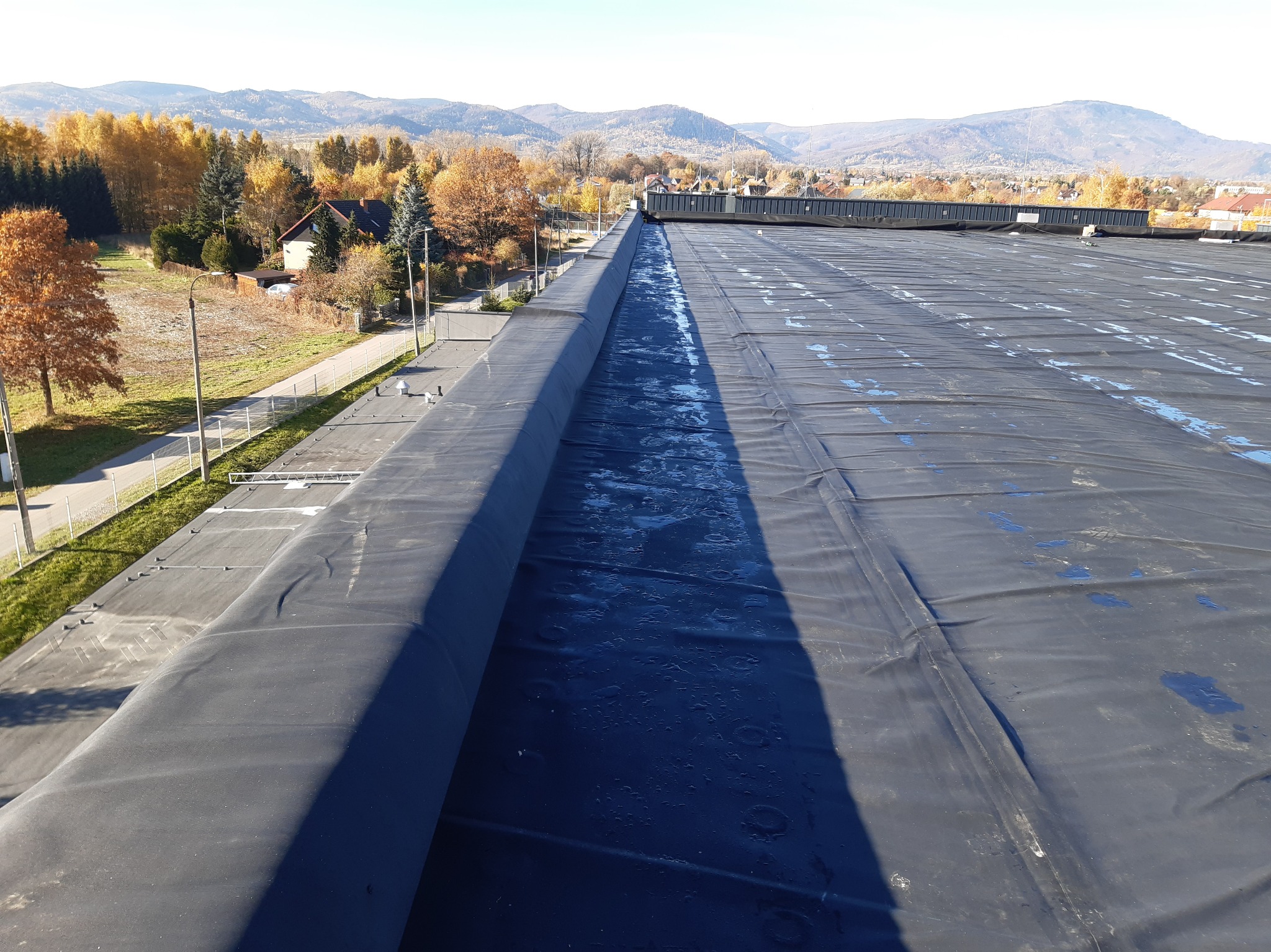 Widok z góry na płaski dach pokryty czarną membraną, z widocznymi śladami wilgoci i odbiciami światła, w tle krajobraz zalesionych wzgórz w jesiennej kolorystyce pod błękitnym niebem.