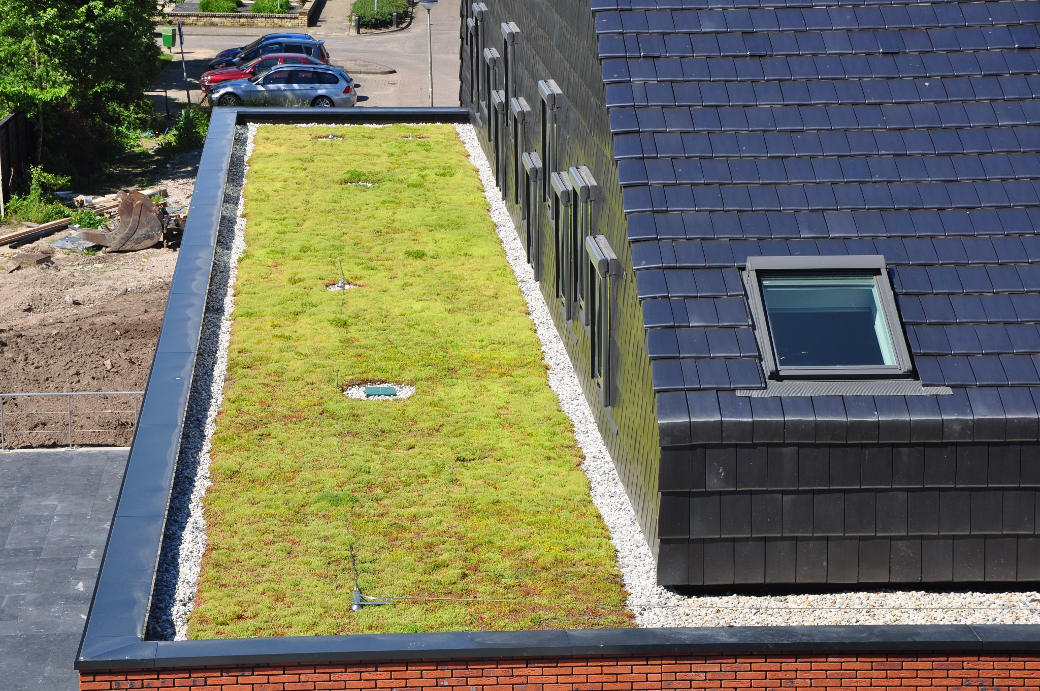 Widok z góry na zielony dach z roślinnością, obok dach pokryty czarną dachówką z oknem dachowym, oddzielone pasem białego żwiru.