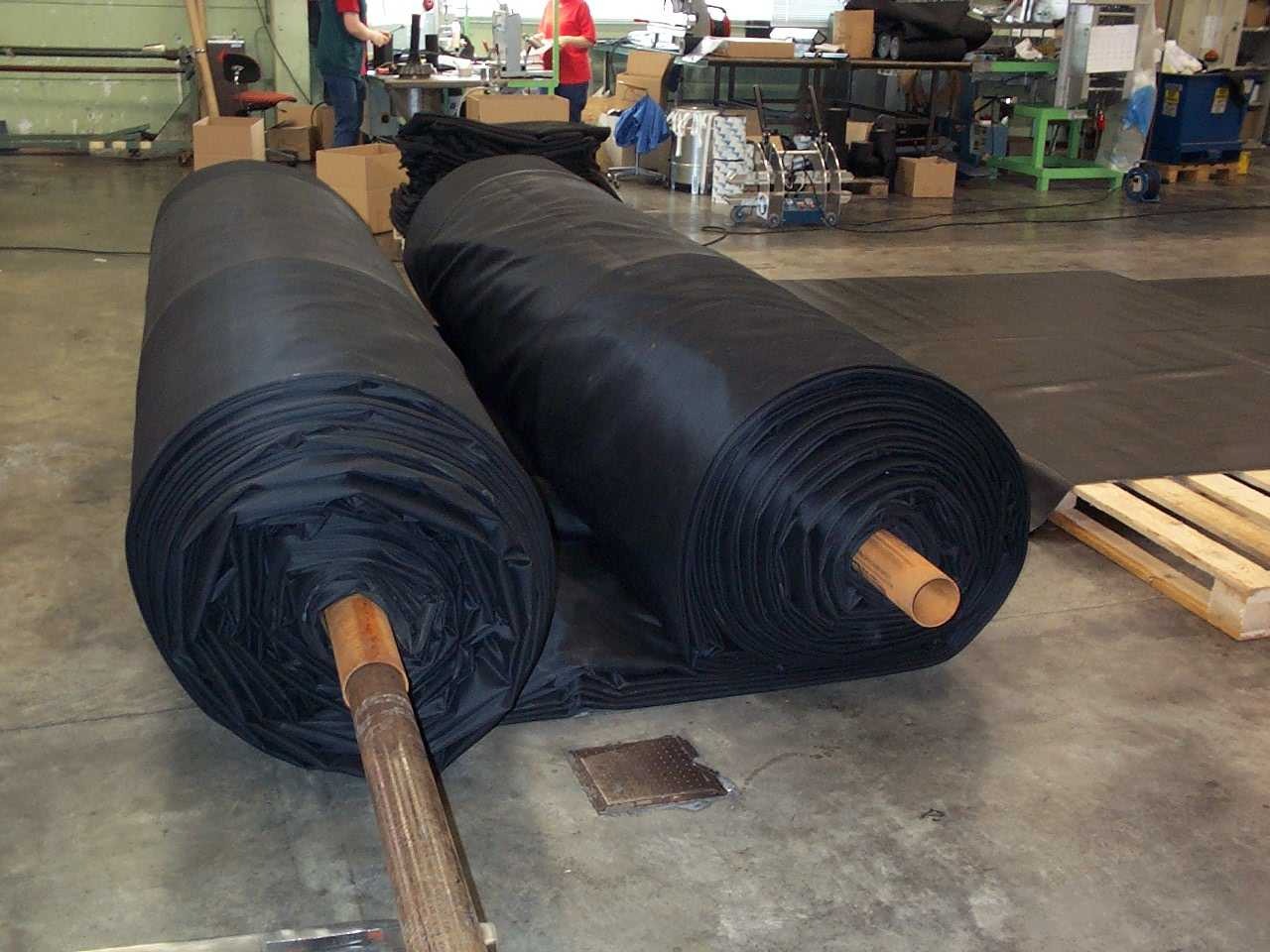 Dwa duże zwoje czarnej membrany dachowej na drewnianych rdzeniach, rozłożony fragment membrany, paleta i wyposażenie warsztatowe w tle.