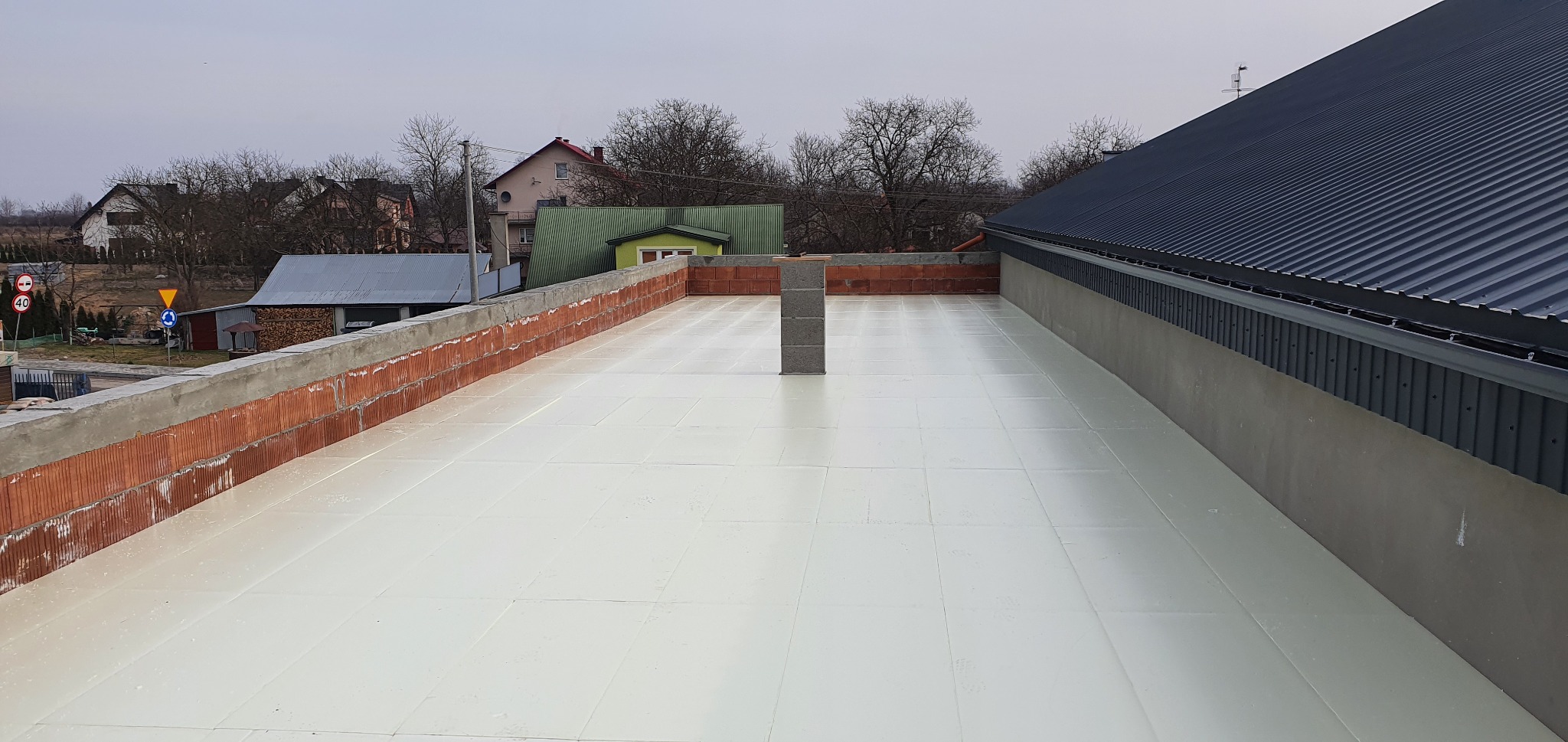 Przygotowana powierzchnia dachu pokryta białym materiałem izolacyjnym, obrzeże z cegieł i szara blacha dachowa w tle krajobraz wiejski.