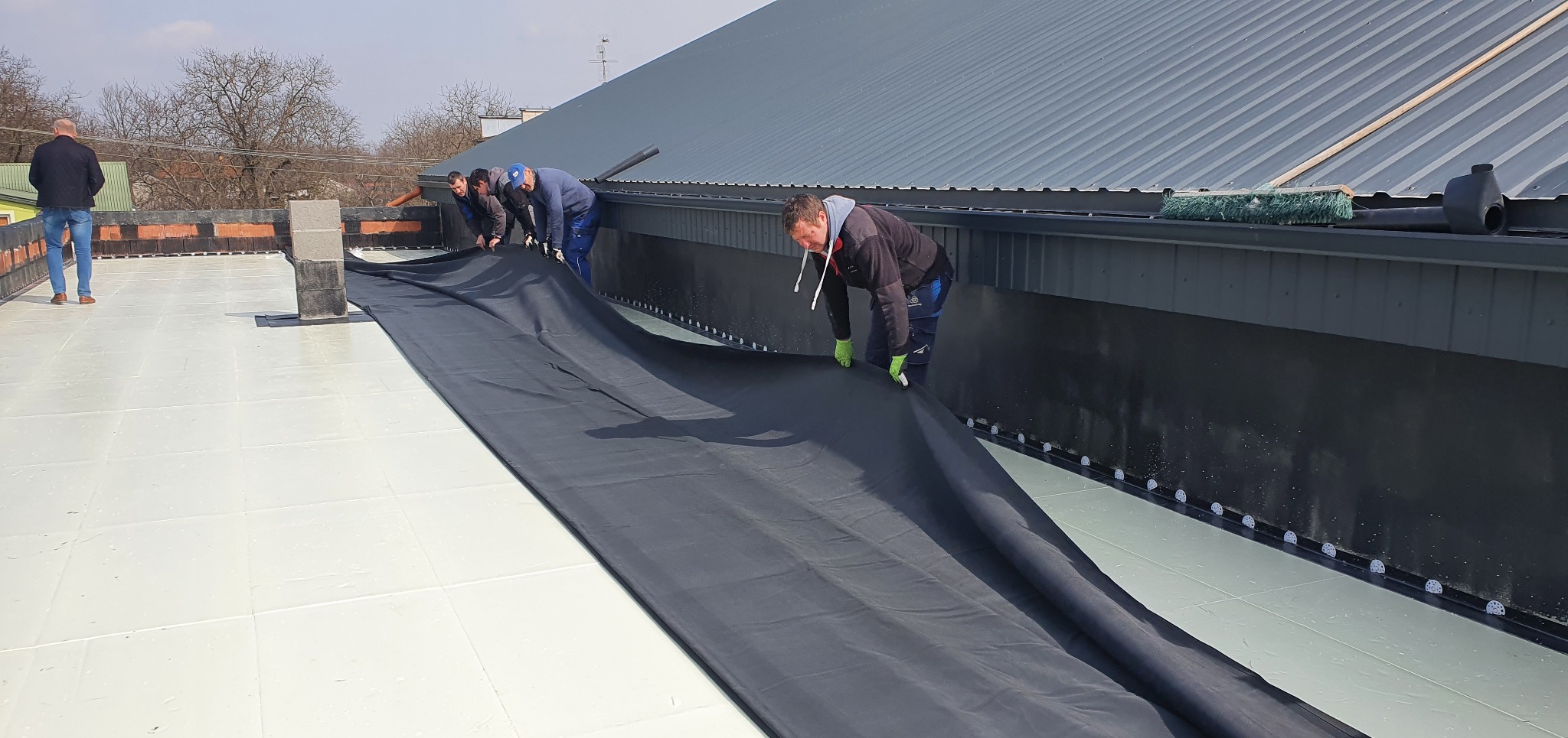 Ekipa dekarzy rozkłada czarną membranę hydroizolacyjną na płaskim dachu z widocznymi elementami mocującymi, w tle blaszany dach i budynki.