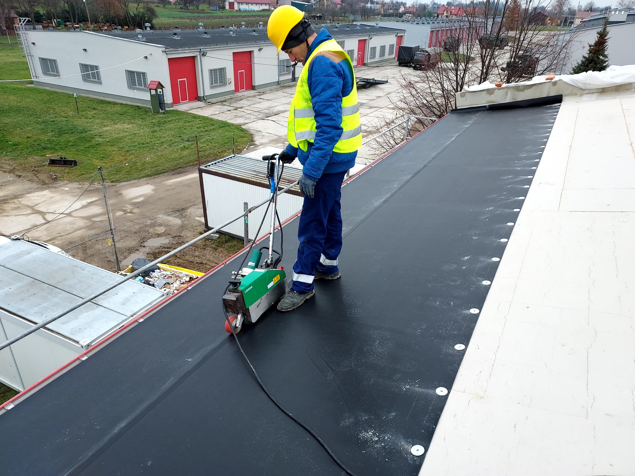 Pracownik w żółtym kasku i kamizelce spawa papę termozgrzewalną na dachu budynku przemysłowego, używając specjalistycznego sprzętu.