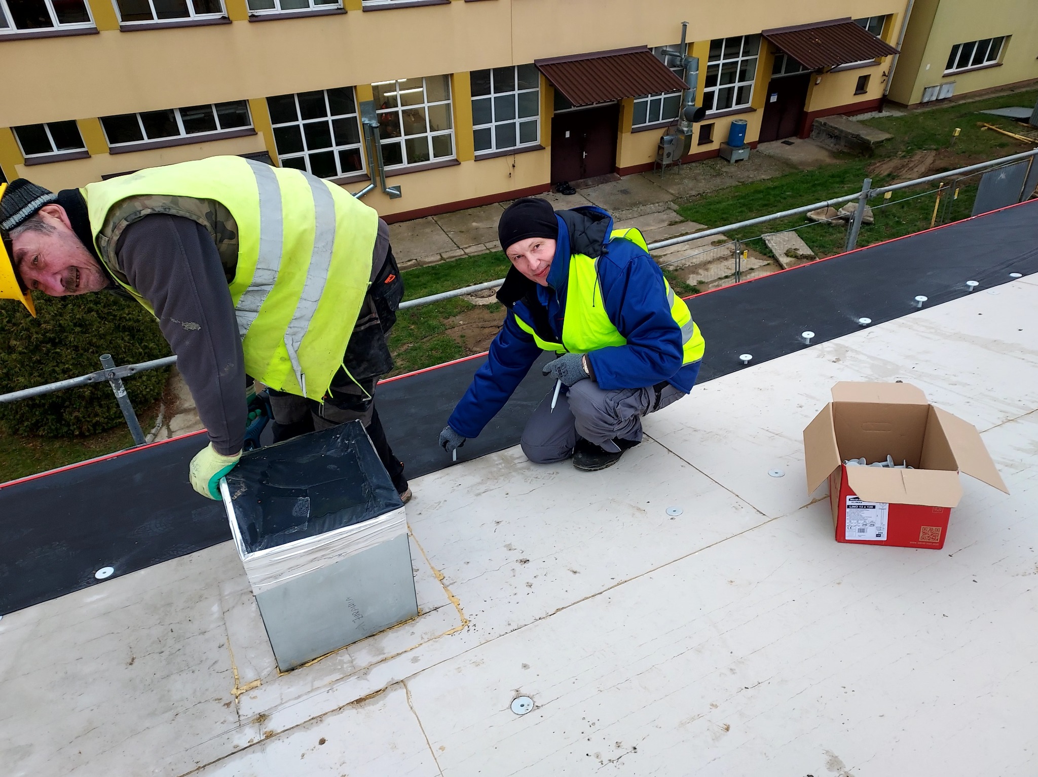 Dwóch dekarzy w ubraniach roboczych na dachu budynku, jeden z wiertarką montuje element wentylacyjny, drugi mierzy i zaznacza punkty mocowania membrany dachowej.