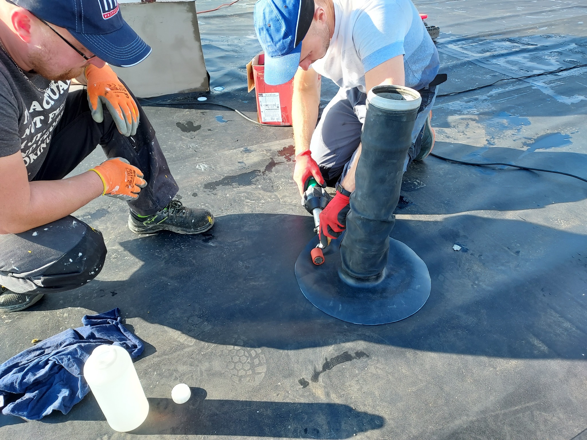 Dwóch dekarzy pracuje na płaskim dachu, uszczelniając przejście rury wentylacyjnej za pomocą zgrzewarki dekarskiej i membrany bitumicznej.