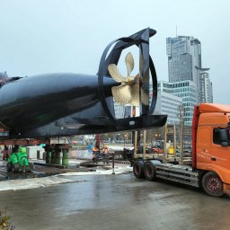 TYMCZASOWE DROGI MODUŁOWE SPÓŁKA Z OGRANICZONĄ ODPOWIEDZIALNOŚCIĄ - Transport łodzi podwodnej ORP Sokół na platformie ciężarówki Volvo, zabezpieczonej systemem ALU Deck HD, z widoczną śrubą okrętową i budynkami w tle.