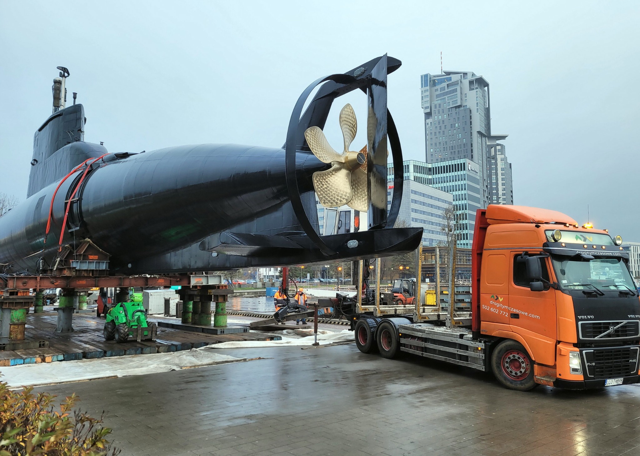 Transport łodzi podwodnej ORP Sokół na platformie ciężarówki Volvo, zabezpieczonej systemem ALU Deck HD, z widoczną śrubą okrętową i budynkami w tle.