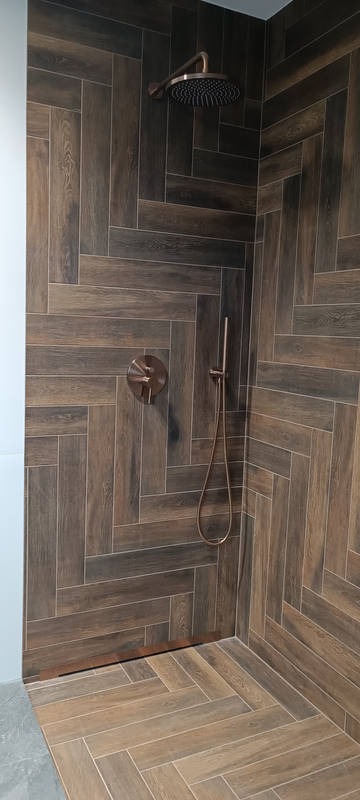 Kabina prysznicowa z drewnopodobnymi płytkami ułożonymi w jodełkę. Armatura w kolorze miedzianym. Ściek liniowy przy ścianie. Nowoczesny design.