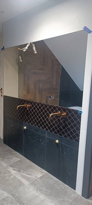 Wykończona łazienka: ciemne płytki imitujące marmur, mozaika nad umywalkami, złote baterie, duże lustro. Widoczne elementy instalacji elektrycznej.