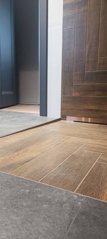 Ukończona podłoga w łazience z brązowych płytek ułożonych w jodełkę i szarych płytek przy progu. Widoczny fragment ściany z drewnopodobnych płytek.