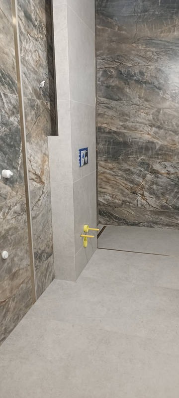 Wykończona łazienka z płytkami imitującymi marmur na ścianach i szarymi płytkami na podłodze. Widoczne przyłącza do armatury i zabudowa stelaża WC.