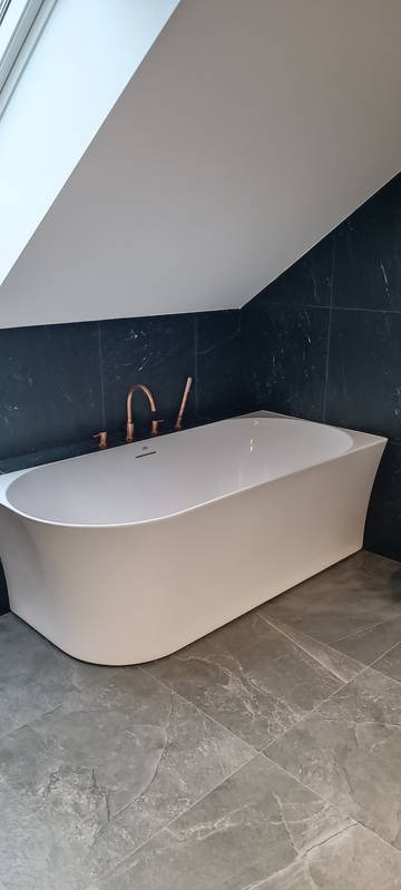 Nowoczesna, biała wanna wolnostojąca z miedzianą armaturą na tle ciemnych, marmurkowych płytek i szarej podłogi w łazience na poddaszu.
