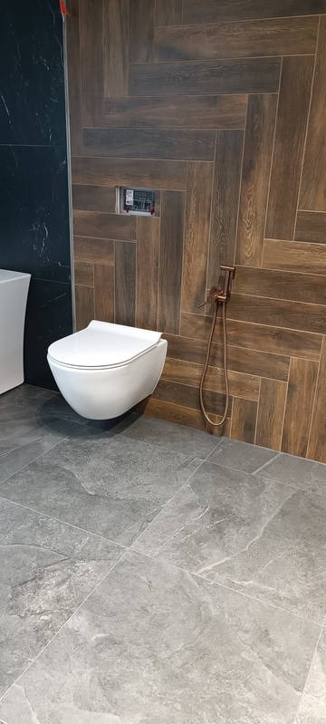 Nowoczesna łazienka z drewnopodobnymi płytkami ułożonymi w jodełkę na ścianie, szara podłoga i podwieszana toaleta z bidetką w kolorze miedzi.