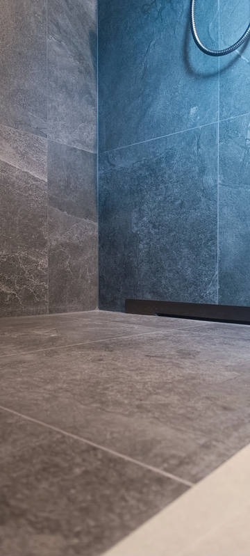 Nowoczesna kabina prysznicowa z szarymi płytkami imitującymi kamień na ścianach i podłodze oraz czarnym odpływem liniowym. Widoczny fragment węża prysznicowego.