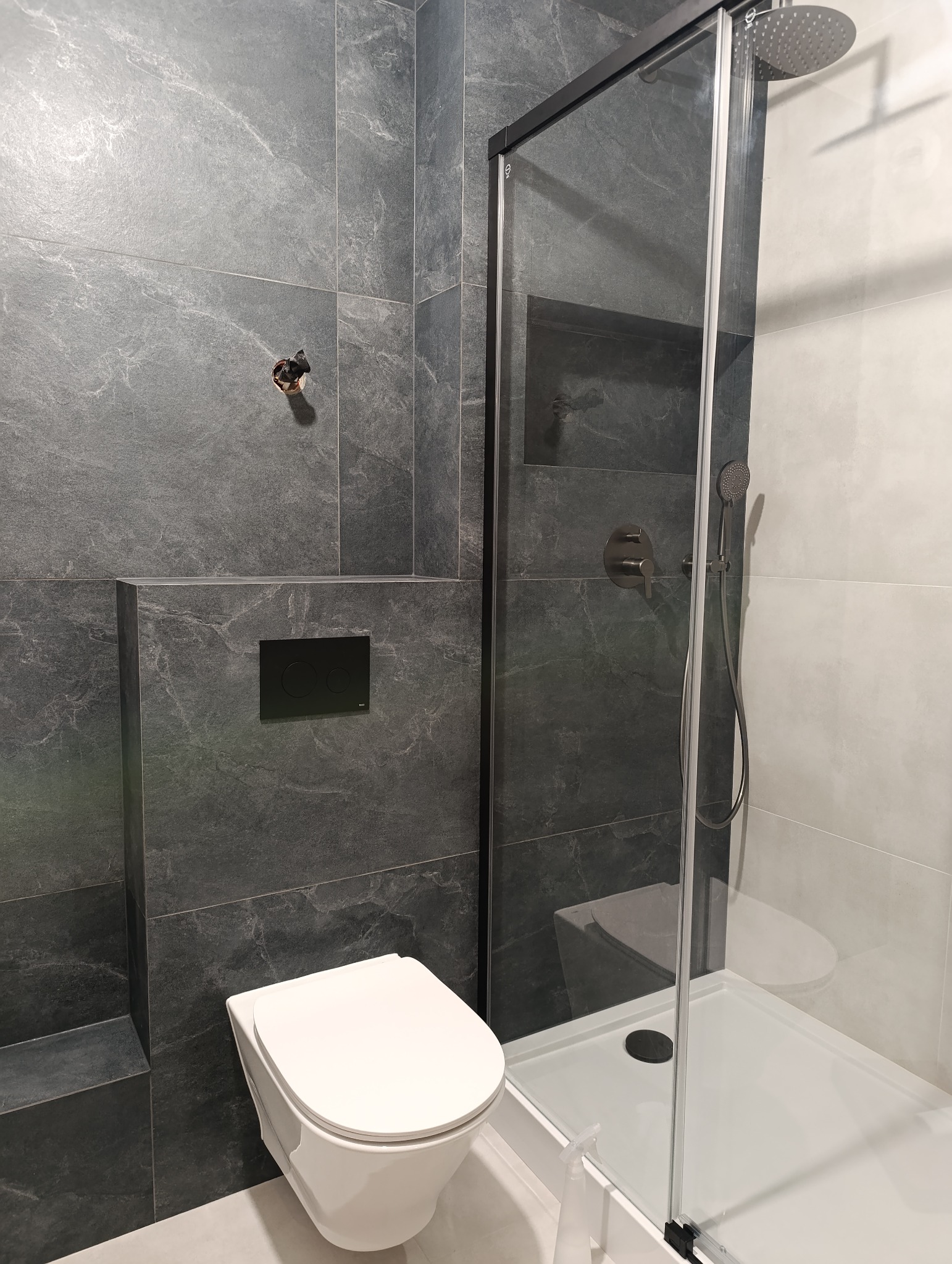 Nowoczesna łazienka z szarymi płytkami imitującymi kamień, kabiną prysznicową ze szklanymi drzwiami i podwieszaną toaletą. Widoczny otwór na baterię.