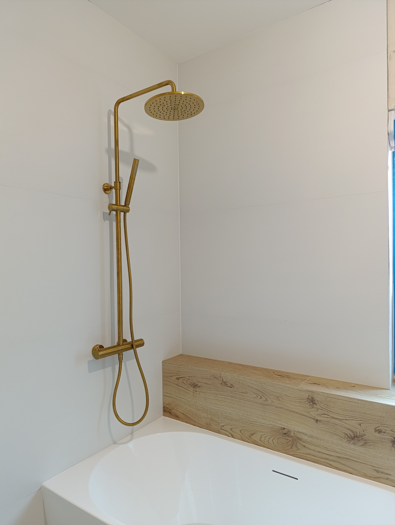 Biała wanna z panelem imitującym drewno i złotą baterią natryskową na tle białych płytek w minimalistycznej łazience.