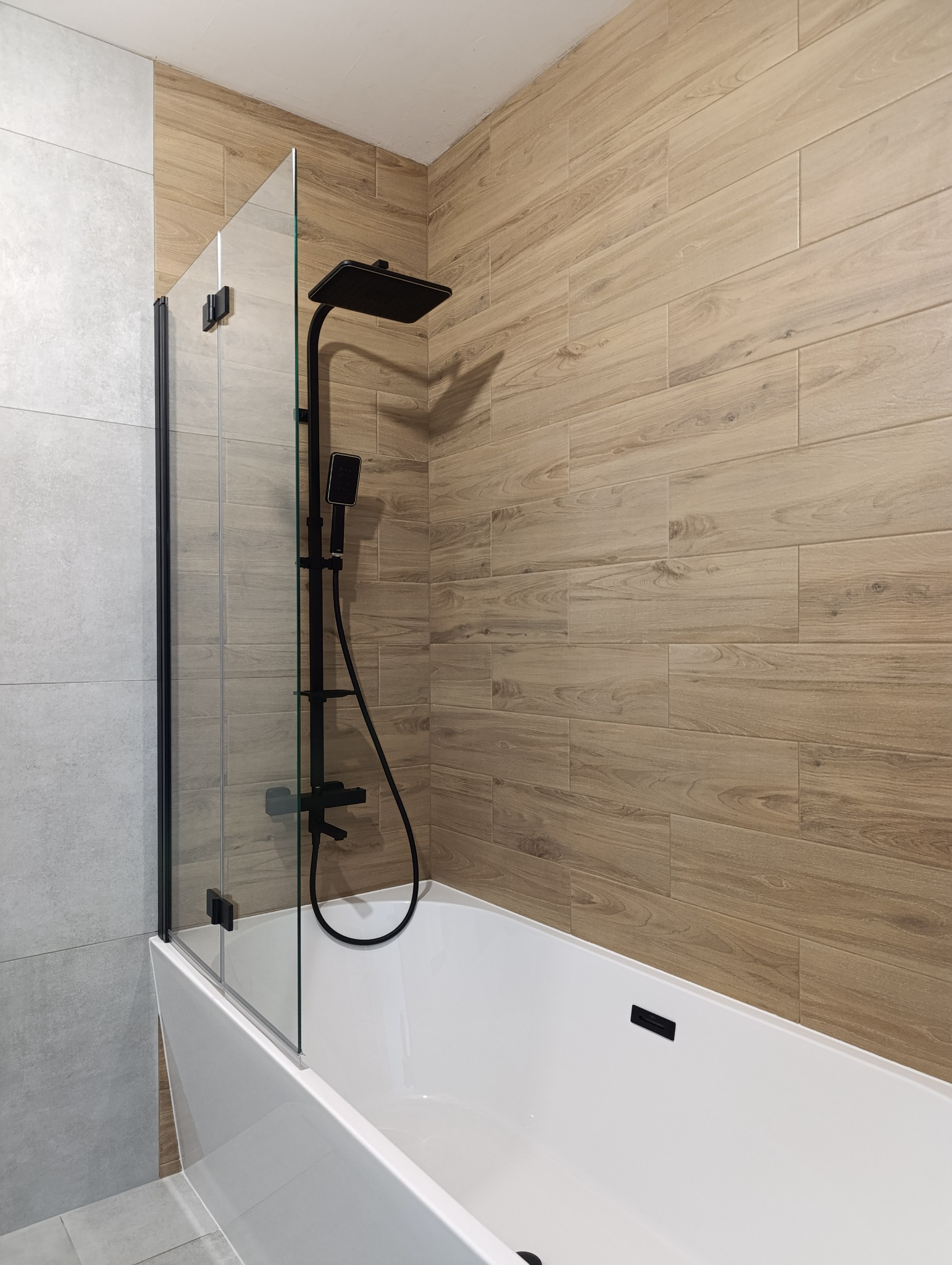 Nowoczesna łazienka po remoncie: biała wanna z czarną armaturą, szklany parawan nawannowy i płytki imitujące drewno na ścianie, jasne płytki na podłodze.