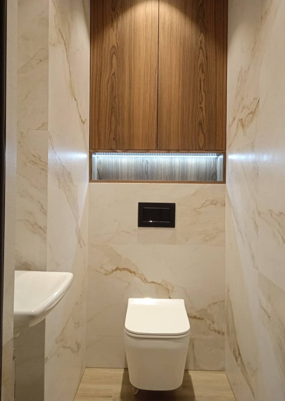 Nowoczesna łazienka z marmurowymi płytkami, drewnianą szafką z oświetleniem LED, wiszącą miską WC i umywalką. Minimalistyczny design, ciepłe oświetlenie.