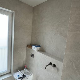 Sergio - Montaż podwieszanej toalety w łazience wykończonej szarymi płytkami imitującymi marmur, widoczne narzędzia i chemia budowlana, czarna bateria umywalkowa podtynkowa.