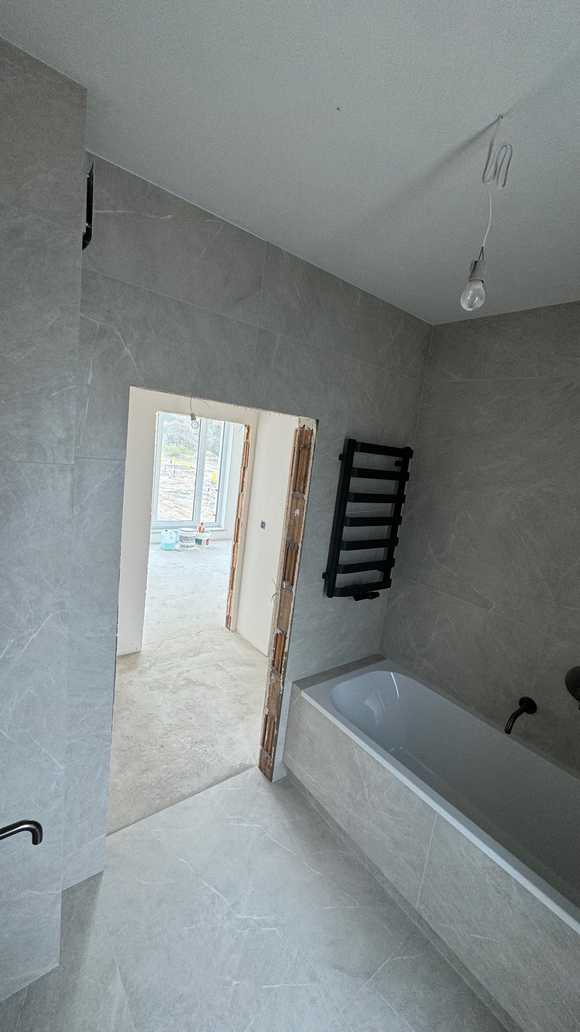 Łazienka w trakcie wykańczania, szare płytki imitujące marmur na ścianach i podłodze, widoczny grzejnik drabinkowy, wanna w zabudowie, surowa oprawa żarówki zwisająca z sufitu, przez otwarte drzwi...