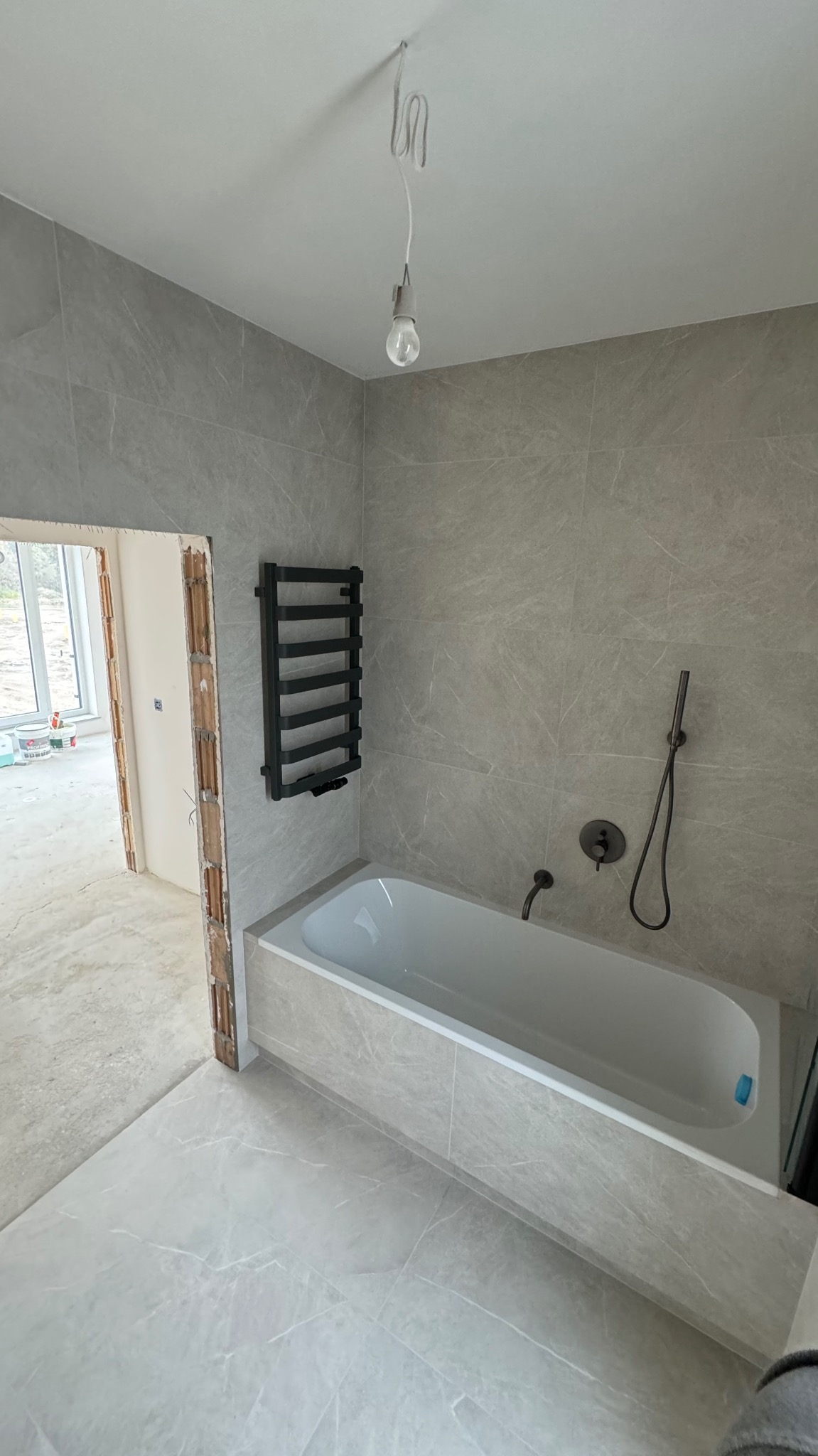 Nowoczesna łazienka w odcieniach szarości z marmurkowymi płytkami, czarnym grzejnikiem drabinkowym i minimalistyczną armaturą, widok na sąsiednie pomieszczenie w trakcie wykończenia.