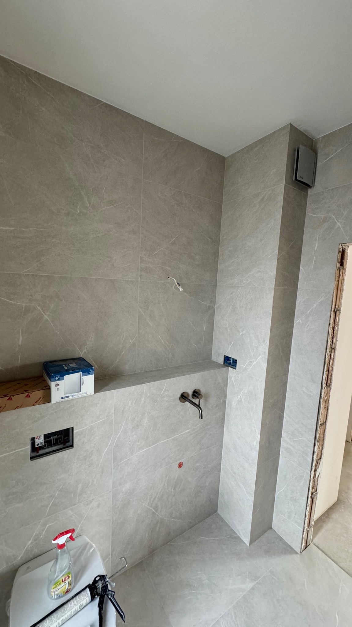 Wykończona łazienka z szarymi płytkami imitującymi marmur, widoczna bateria umywalkowa podtynkowa, częściowo zamontowana toaleta z silikonem i środkiem czyszczącym obok, wentylator sufitowy...