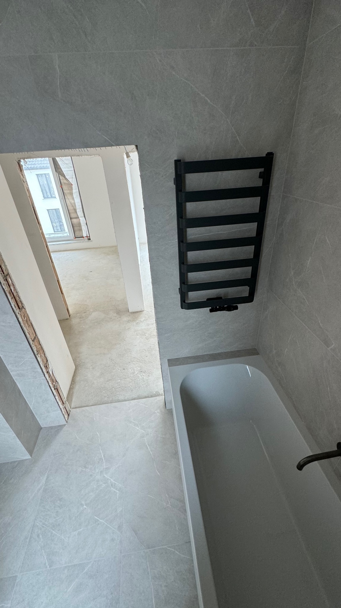 Wykończona łazienka w szarościach z wanną i czarnym grzejnikiem drabinkowym, widok na remontowane pomieszczenie obok.