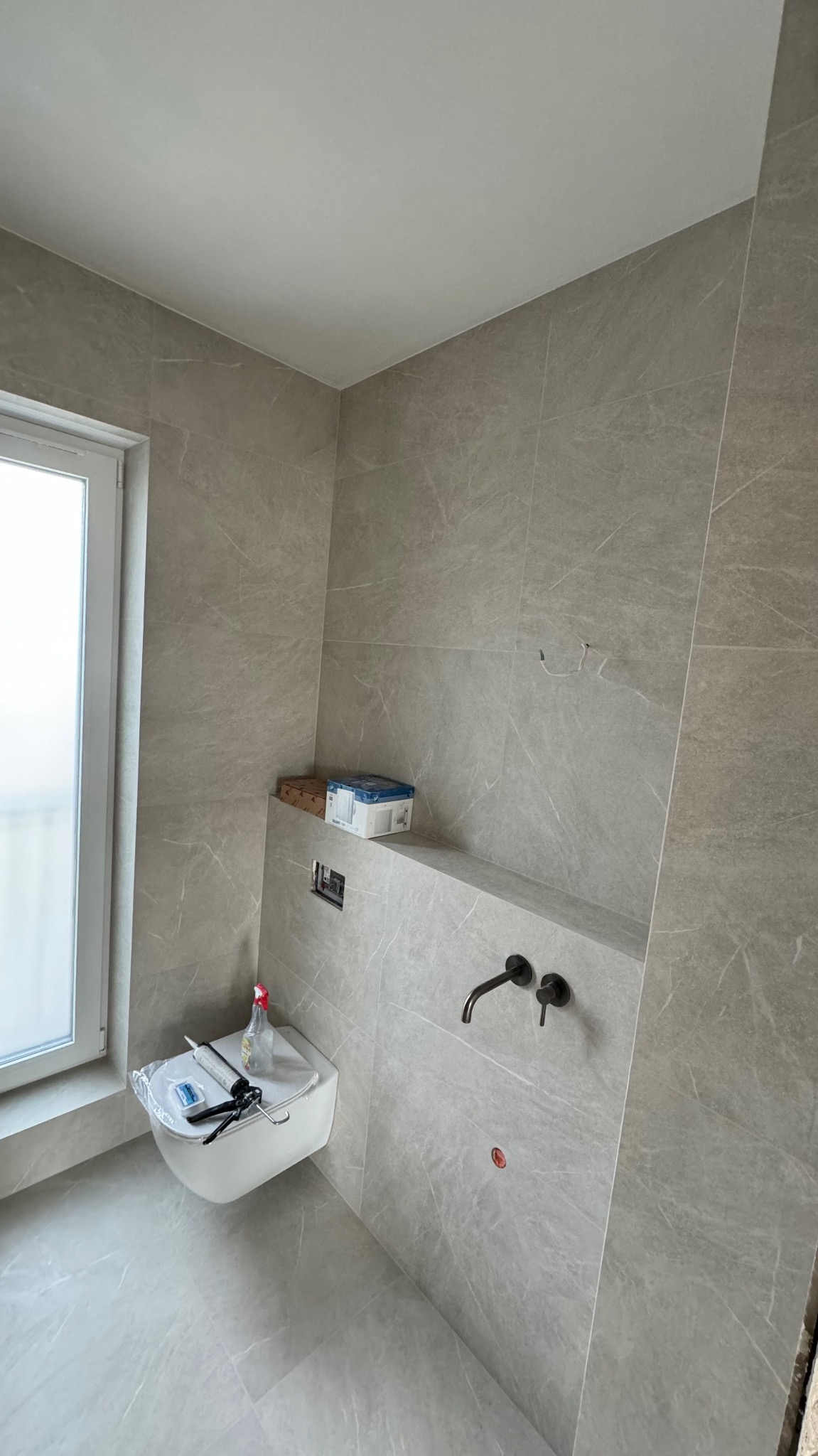 Montaż podwieszanej toalety w łazience wykończonej szarymi płytkami imitującymi marmur, widoczne narzędzia i chemia budowlana, czarna bateria umywalkowa podtynkowa.