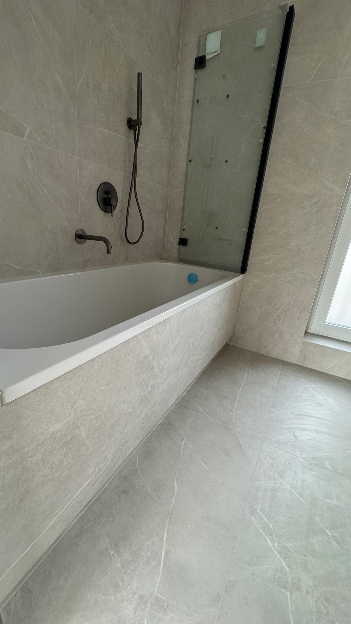 Nowoczesna łazienka wykończona w szarych płytkach imitujących marmur, z białą wanną, czarną armaturą i szklanym parawanem nawannowym z czarną ramą, uzupełniona niebieską zabawką w wannie.