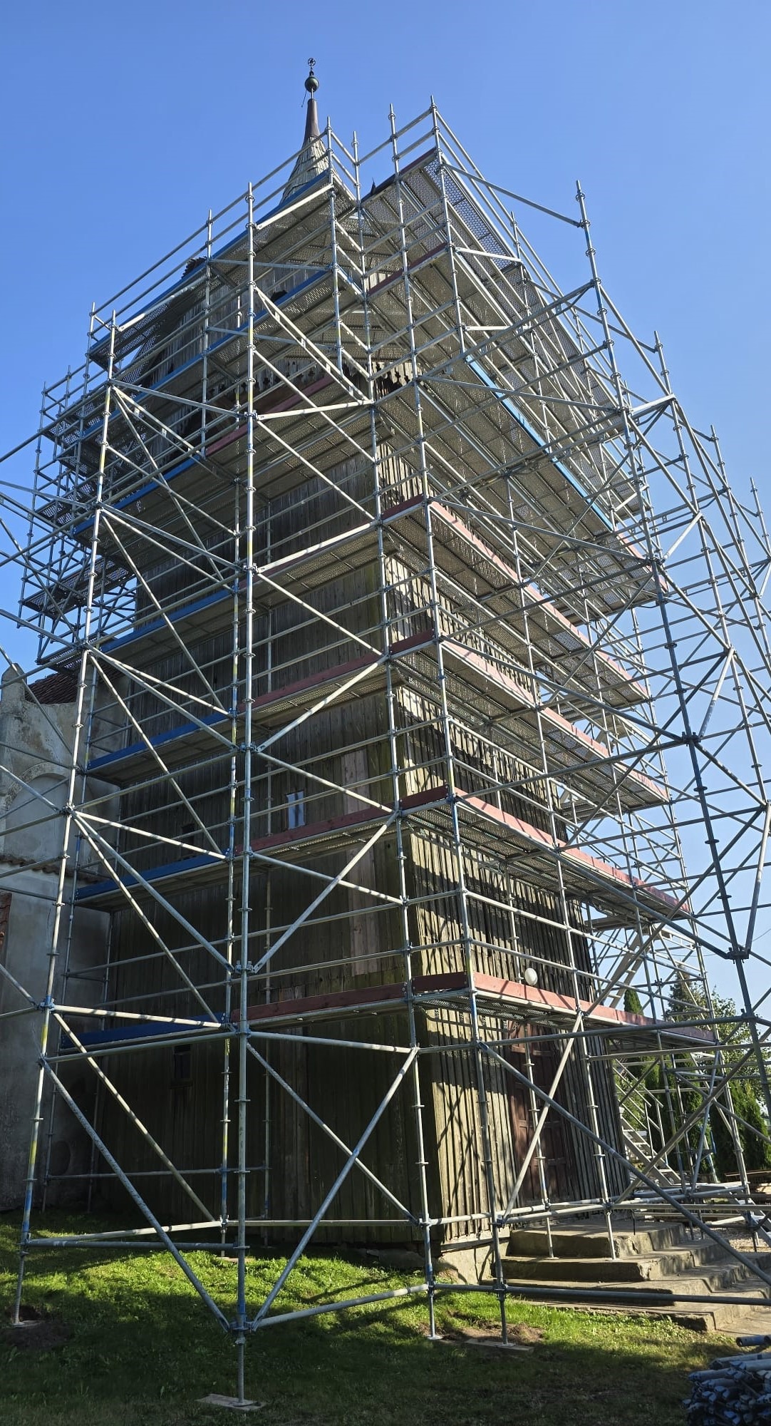 Drewniany kościół w rusztowaniach, widok z dołu na szczyt wieży, błękitne niebo w tle.