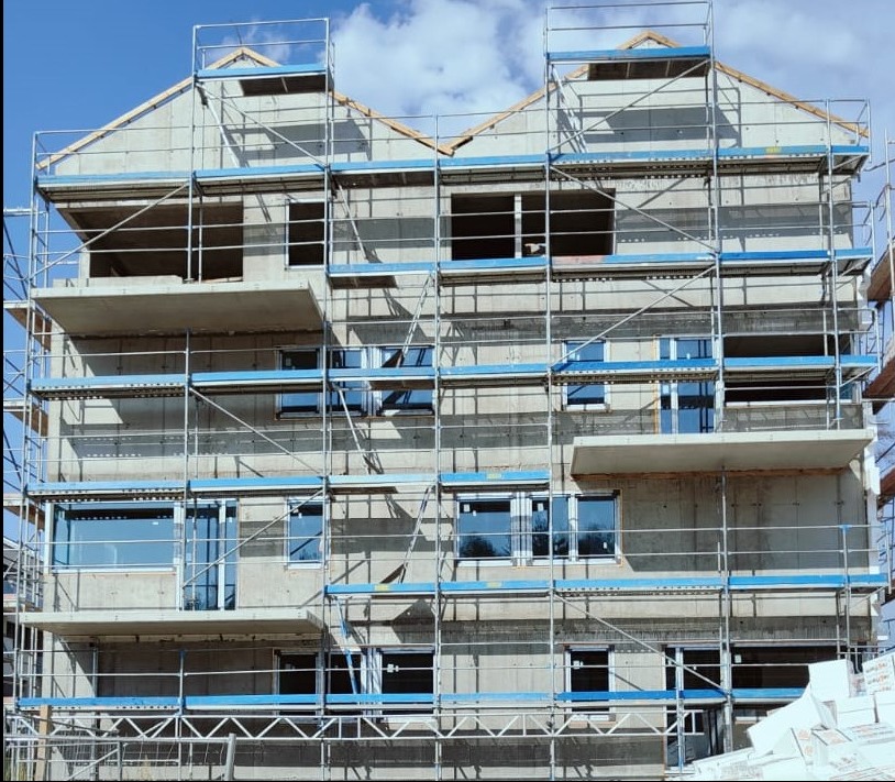 Surowy budynek wielorodzinny w Olsztynie w trakcie budowy, otoczony rusztowaniami, z częściowo wstawionymi oknami, widoczne niebieskie osłony rusztowań.