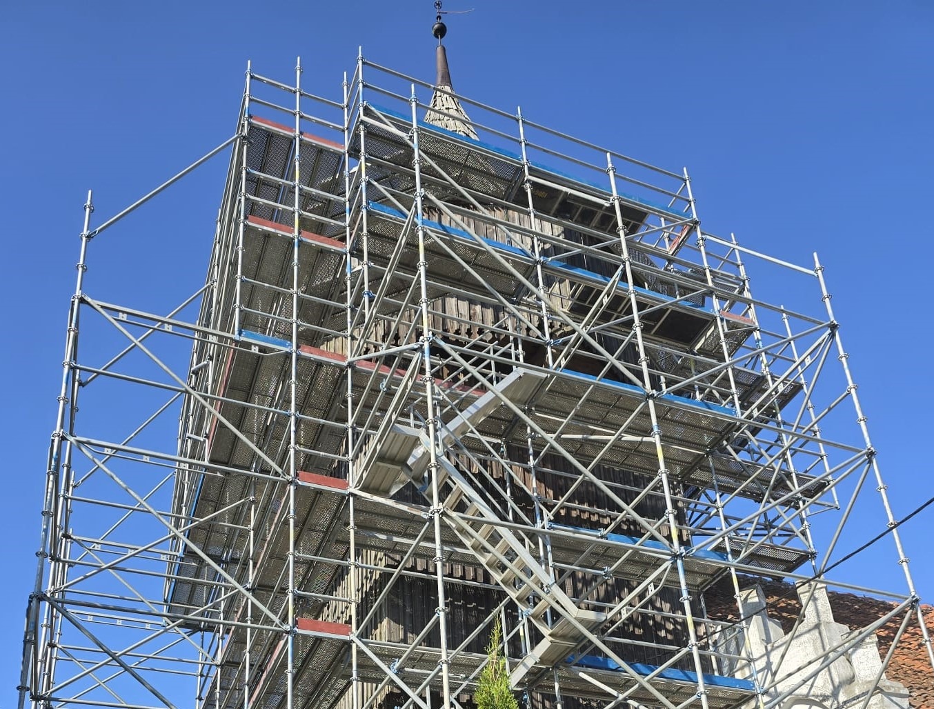 Wysoka wieża kościoła w trakcie renowacji, otoczona rusztowaniem z metalowych rur na tle błękitnego nieba.