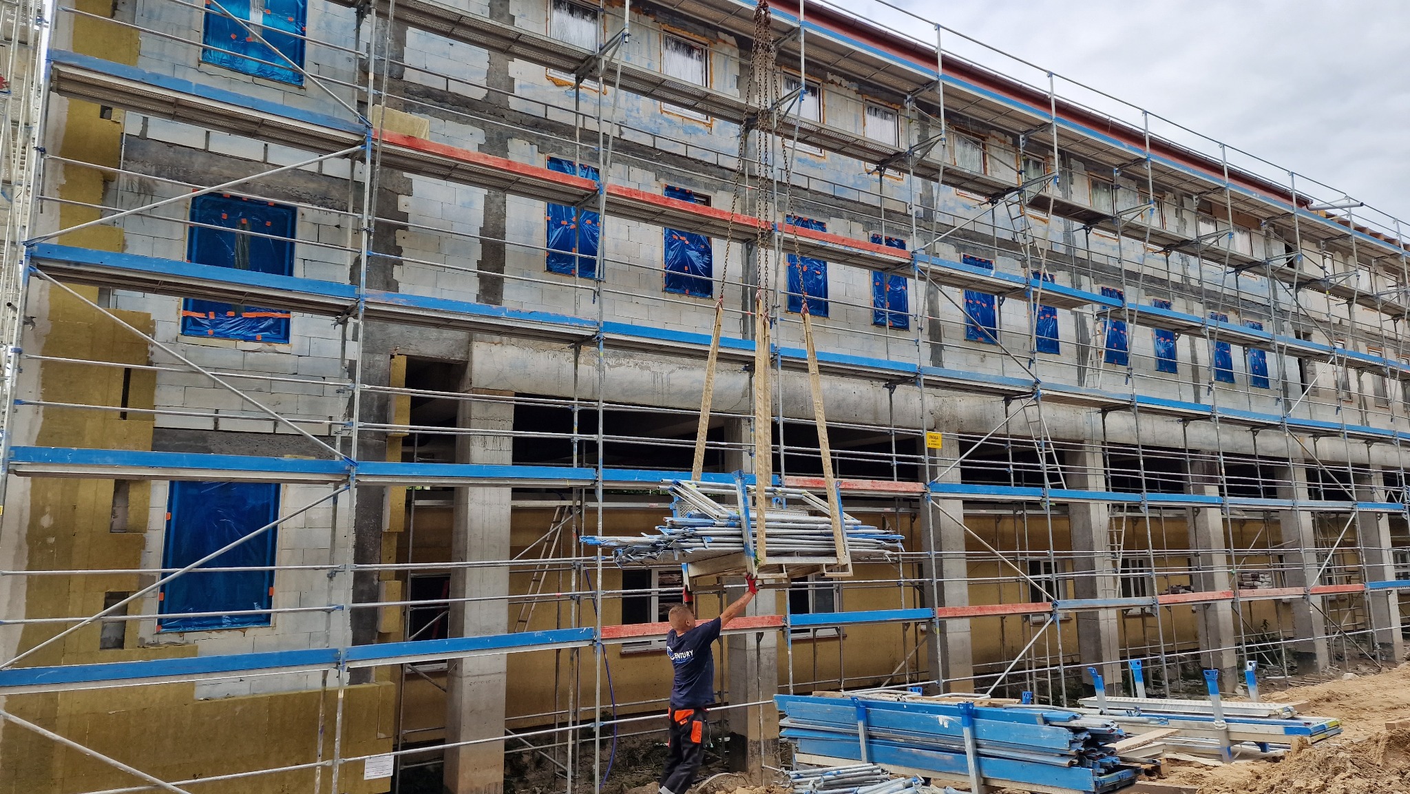 Budynek w trakcie budowy z rusztowaniem, okna zabezpieczone folią, pracownik w czerwonych rękawicach podnosi elementy rusztowania za pomocą dźwigu linowego.