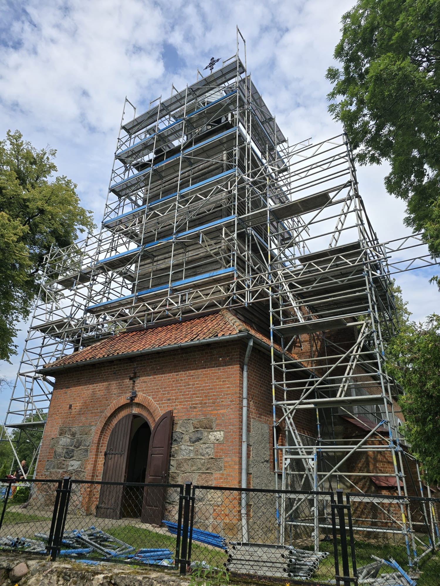 Kościół w trakcie renowacji, otoczony rusztowaniami, widok z dołu, częściowo ogrodzony siatką.
