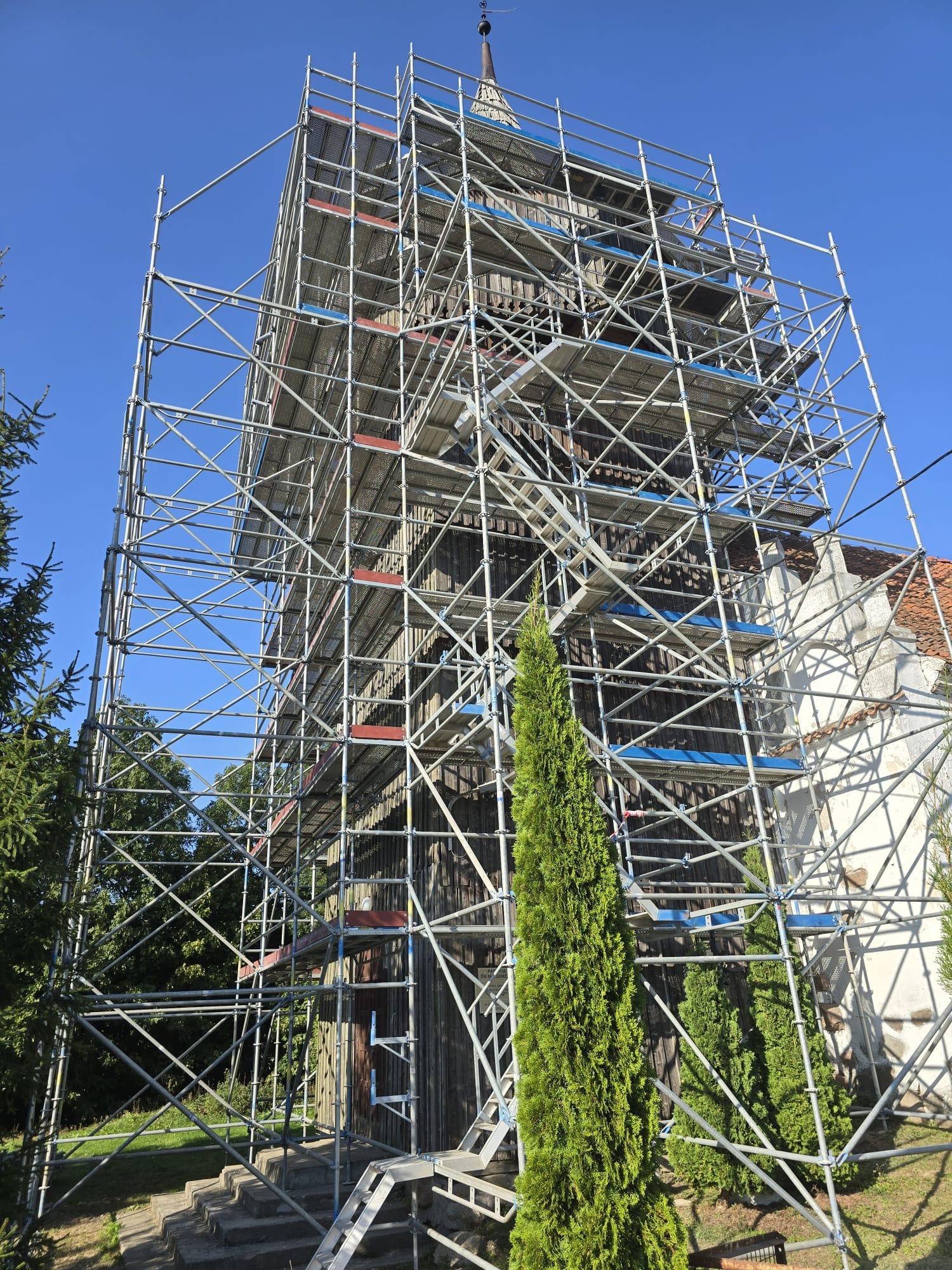 Drewniany kościół w trakcie renowacji, otoczony rusztowaniem, widok z dołu na tle błękitnego nieba.