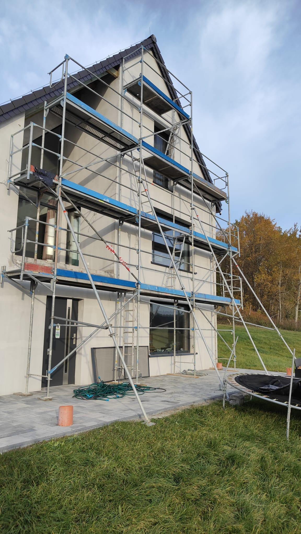 Dwukondygnacyjny dom jednorodzinny w trakcie prac elewacyjnych, otoczony rusztowaniem z niebieskimi elementami, widoczne narzędzia i materiały budowlane na posesji.