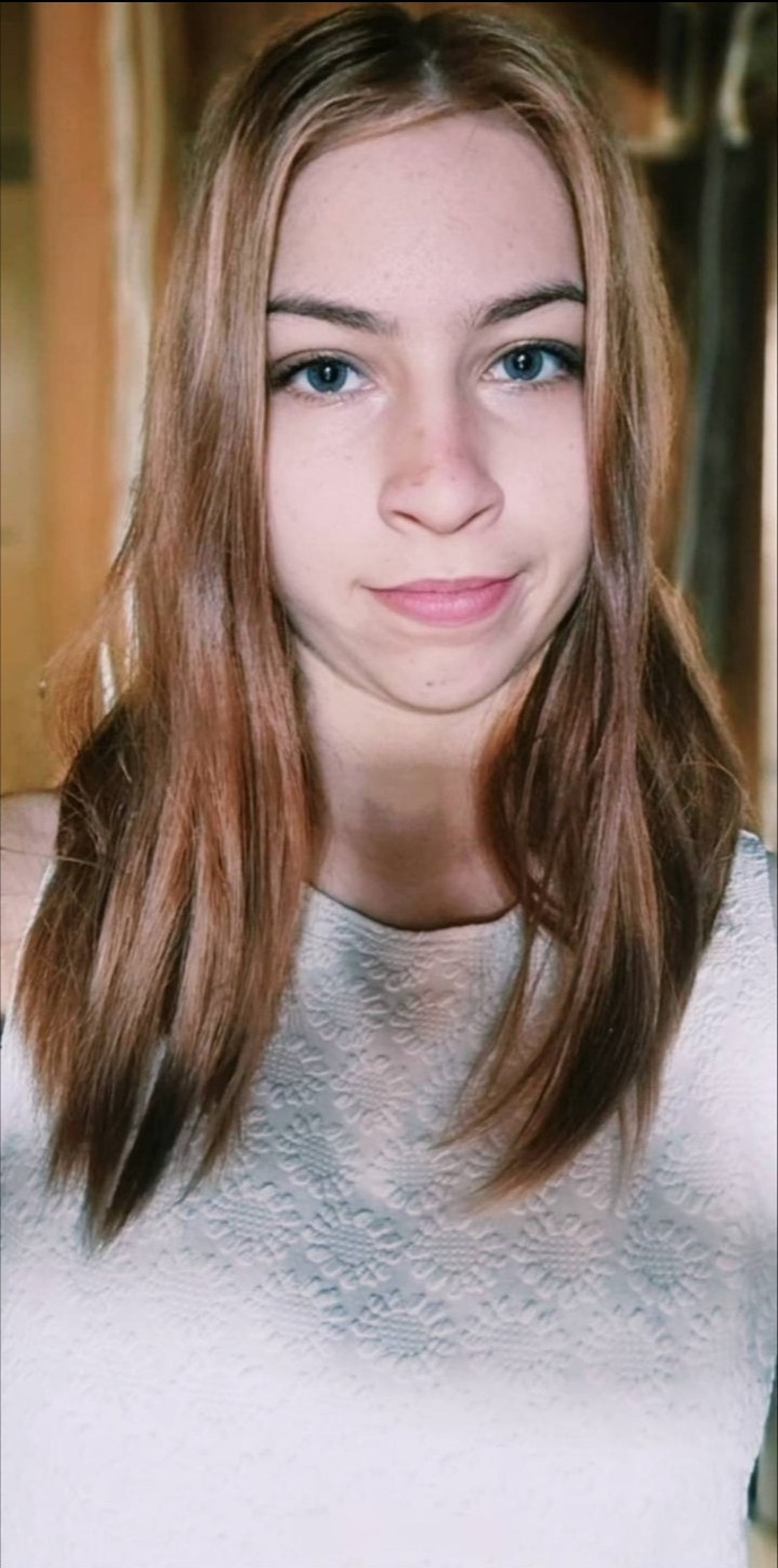 Portret młodej kobiety z długimi, brązowymi włosami i niebieskimi oczami, ubrana w białą bluzkę z wzorem, na neutralnym tle.