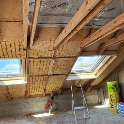 Home Service - Poddasze w trakcie izolacji: widoczne drewniane krokwie, wełna mineralna pomiędzy nimi, dwa okna dachowe, drabina oparta o ścianę, rolki izolacji Isover i płyty gipsowo-kartonowe.