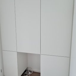 Home Service - Biała zabudowa meblowa z szafkami wiszącymi i dolnymi na brązowej podłodze, z widocznymi kablami w otwartej przestrzeni pod blatem.