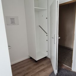 Home Service - Biała szafa z drzwiami, zamontowana w rogu pokoju na drewnianej podłodze, z widocznym szkicem na ścianie i zabezpieczeniem podłogi przed uszkodzeniami.