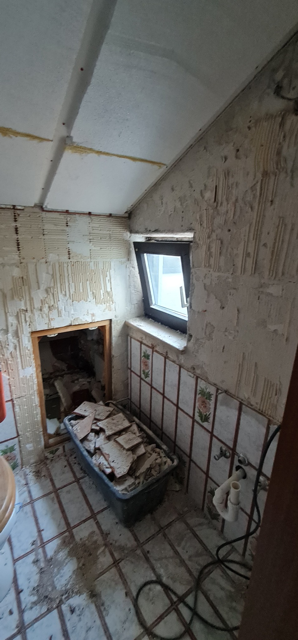 Demontaż płytek w łazience na poddaszu: widoczne ściany z resztkami kleju, okno dachowe, kuweta z odspojonymi płytkami, instalacja wodna.