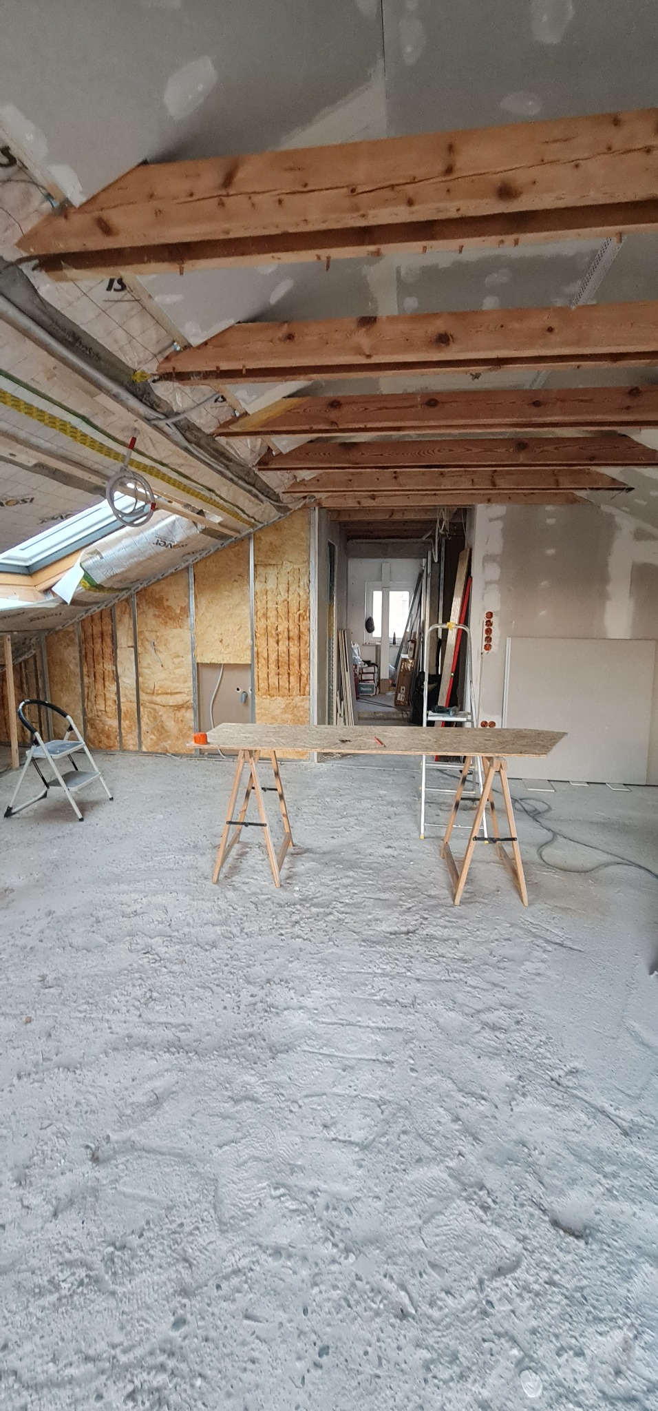 Poddasze w surowym stanie podczas prac wykończeniowych: widoczne ocieplenie ścian wełną mineralną, drewniane belki stropowe, okno dachowe, oraz prowizoryczny stół warsztatowy na kozłach.