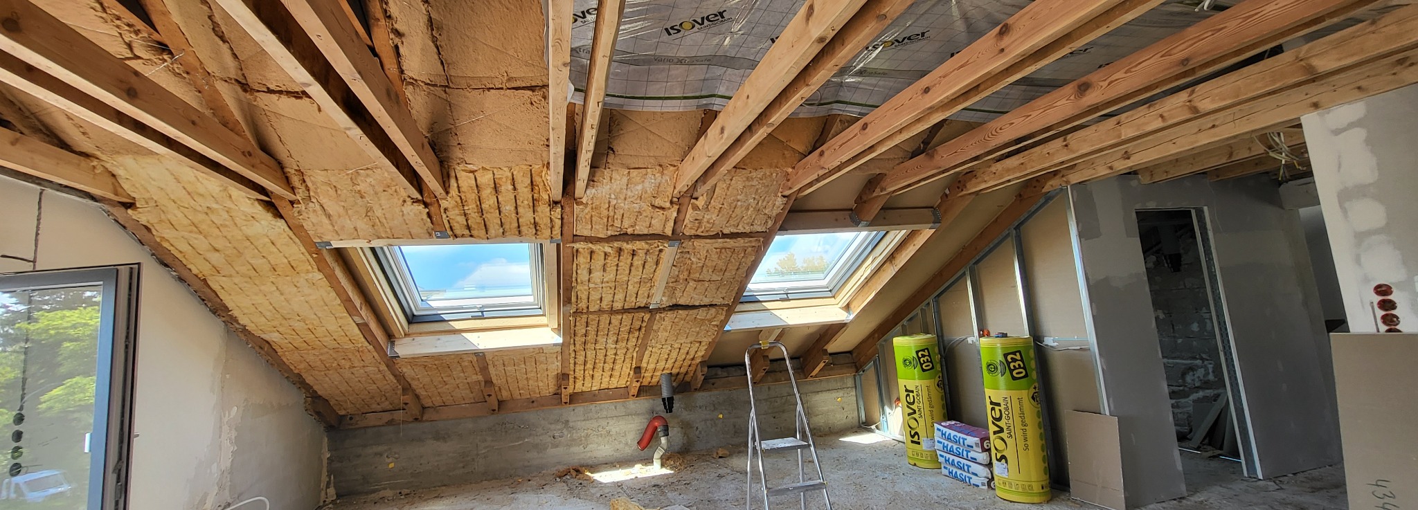 Poddasze w trakcie izolacji: widoczne drewniane krokwie, wełna mineralna pomiędzy nimi, dwa okna dachowe, drabina oparta o ścianę, rolki izolacji Isover i płyty gipsowo-kartonowe.