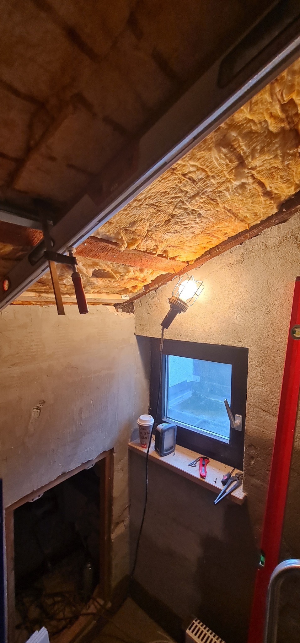 Ocieplenie poddasza wełną mineralną, widoczna lampa robocza, okno z narzędziami na parapecie, poziomica i otwór w ścianie.