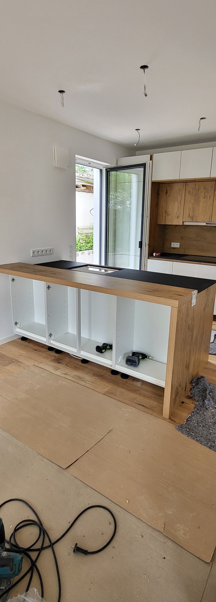 Montaż wyspy kuchennej z blatem w kolorze czarnym i szafkami w kolorze białym i drewnopodobnym, widoczne narzędzia i kable na zabezpieczonej podłodze, w tle otwarte drzwi balkonowe.