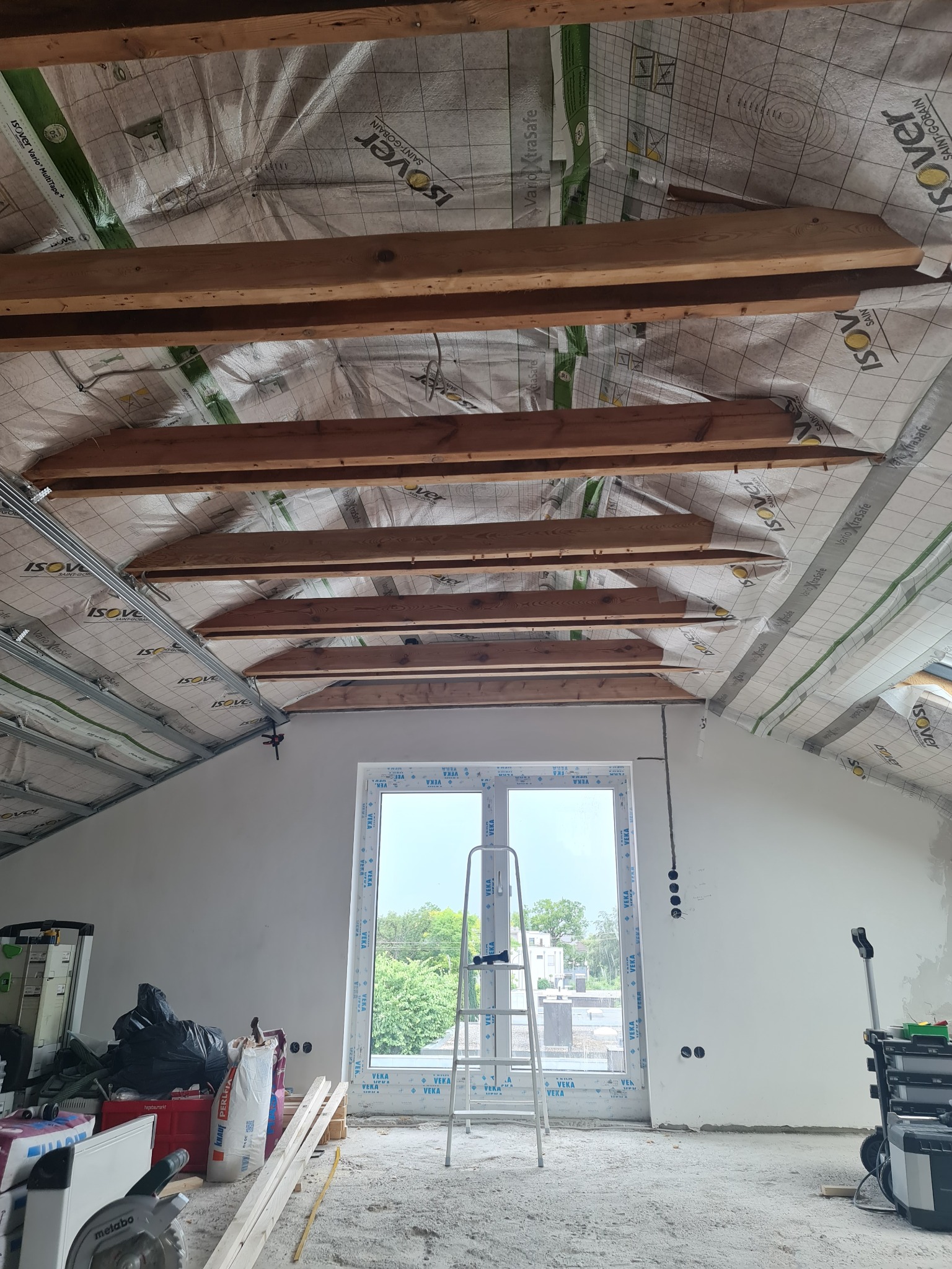 Poddasze w trakcie prac wykończeniowych: ocieplony strop z widocznymi belkami, okno dachowe z drabiną przystawioną do niego, narzędzia i materiały budowlane w nieładzie na podłodze.