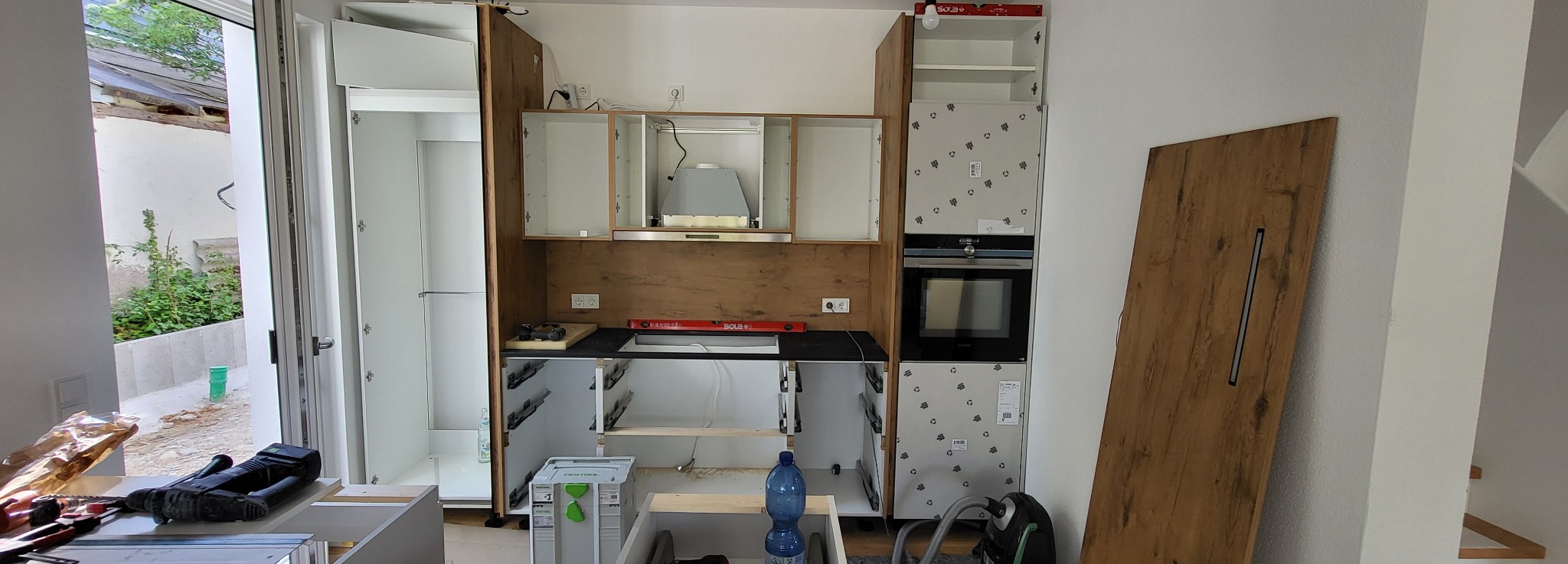 Montaż nowoczesnej kuchni z drewnianymi frontami, widoczne szafki górne bez drzwi, zamontowany okap, piekarnik w zabudowie, poziomica na blacie, narzędzia i butelka wody w tle.