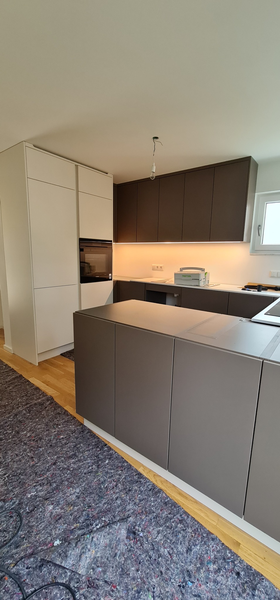Nowoczesna kuchnia w trakcie montażu: szare szafki dolne z wyspą, białe szafki górne z piekarnikiem, surowa żarówka zamiast lampy, podłoga częściowo zabezpieczona folią ochronną.