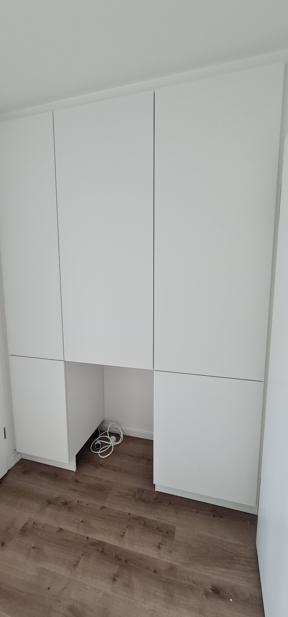 Biała zabudowa meblowa z szafkami wiszącymi i dolnymi na brązowej podłodze, z widocznymi kablami w otwartej przestrzeni pod blatem.