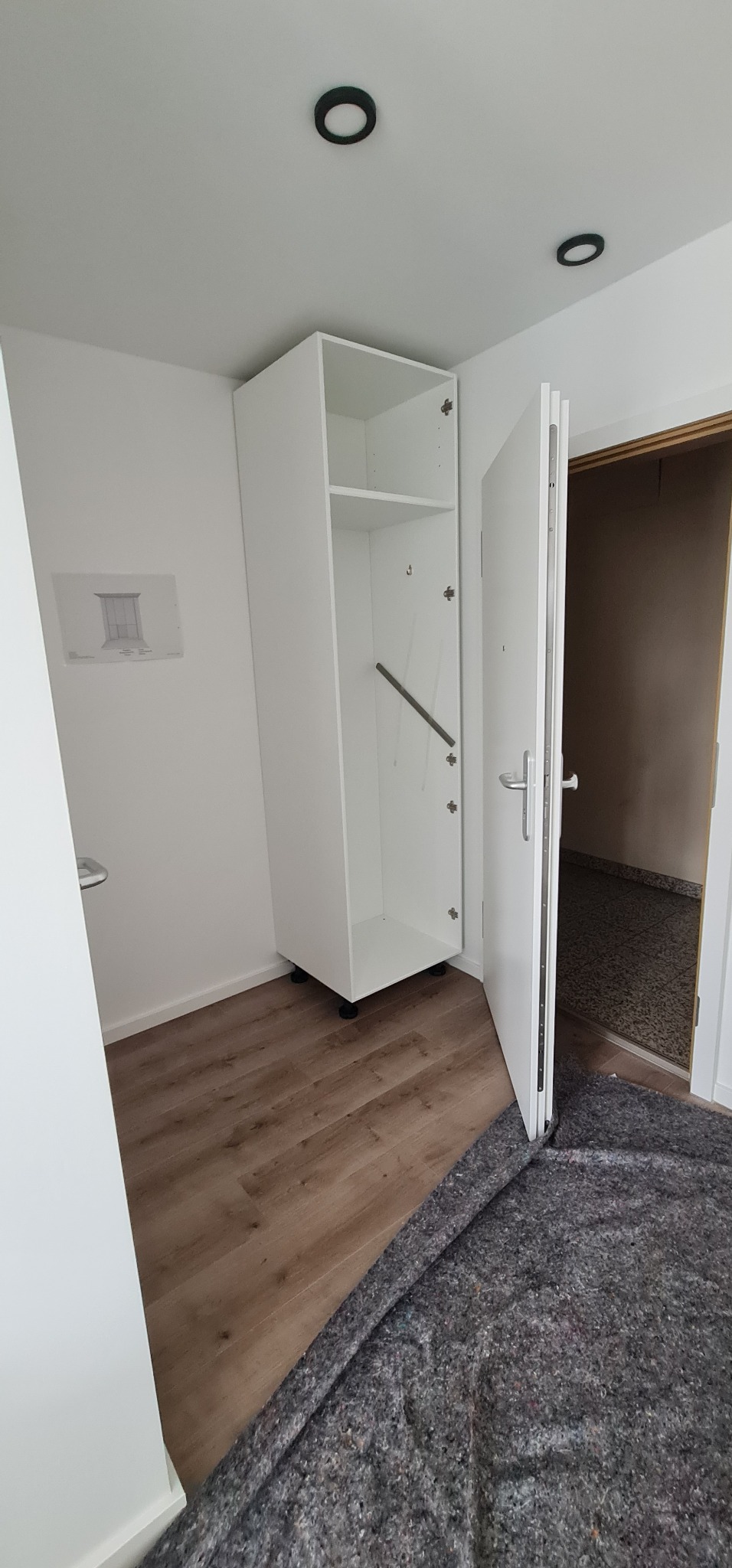 Biała szafa z drzwiami, zamontowana w rogu pokoju na drewnianej podłodze, z widocznym szkicem na ścianie i zabezpieczeniem podłogi przed uszkodzeniami.