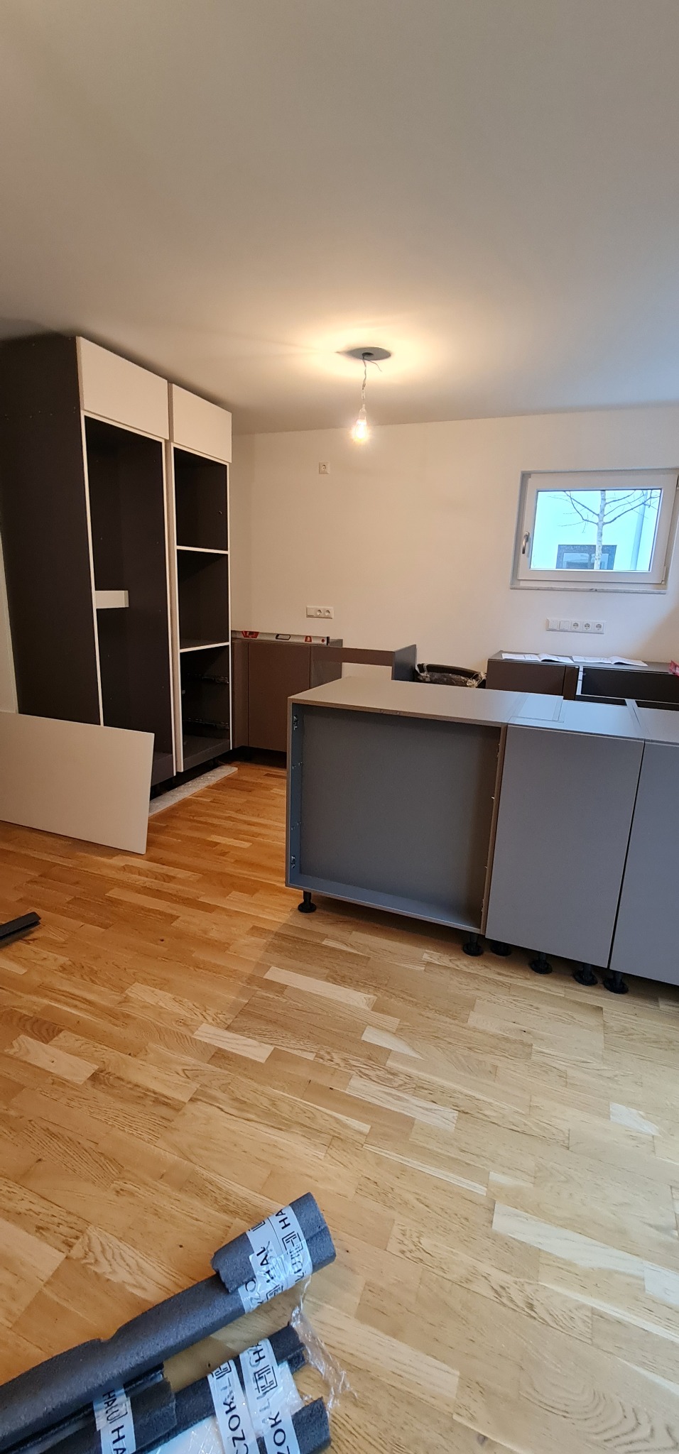 Montaż szarej zabudowy kuchennej z widocznymi elementami szafek i szuflad na regulowanych nóżkach, obok niezmontowana szafa z ciemnymi bokami i białymi frontami, na podłodze parkiet...